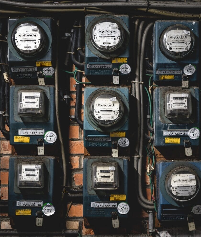 Nine weathered analogue electricity meters mounted in three rows on an old brick wall, each with a glass-domed dial, a padlock, and a numbered tag.