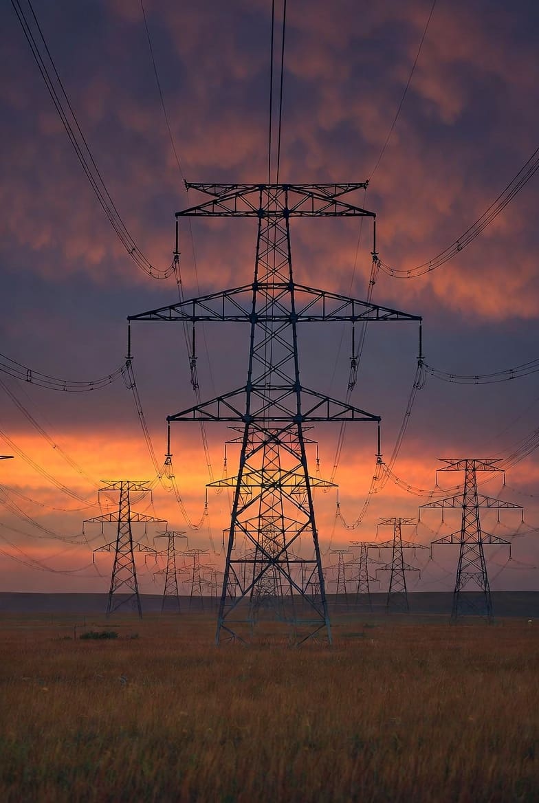 High-voltage transmission towers receding into the horizon at dusk across the Alberta prairie, symbolizing strong, enduring grid infrastructure waiting for future demand.